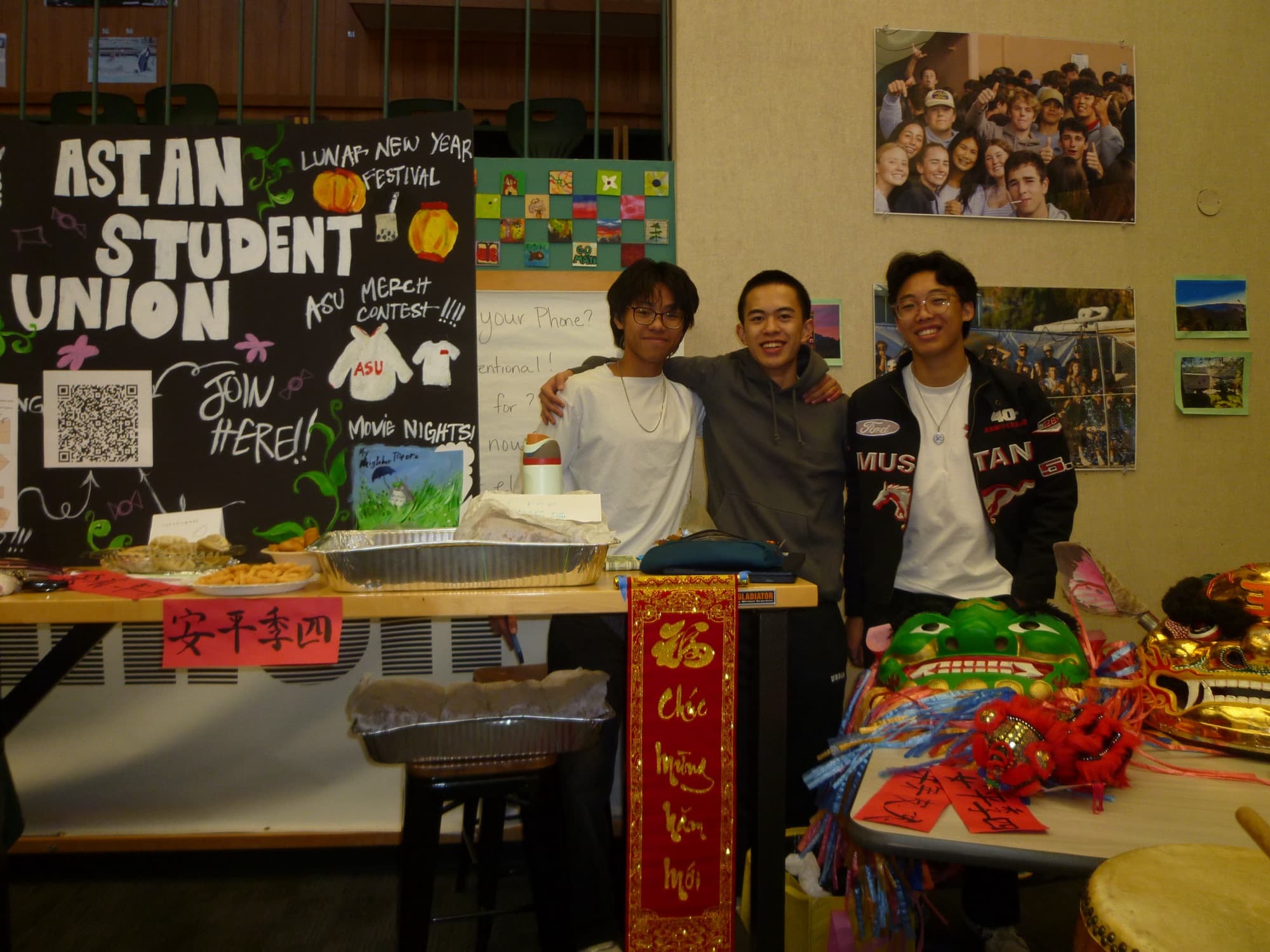 Three students standing behind the Asian Student Union's table, with a hand-lettered poster and dishes laid out in front of them.