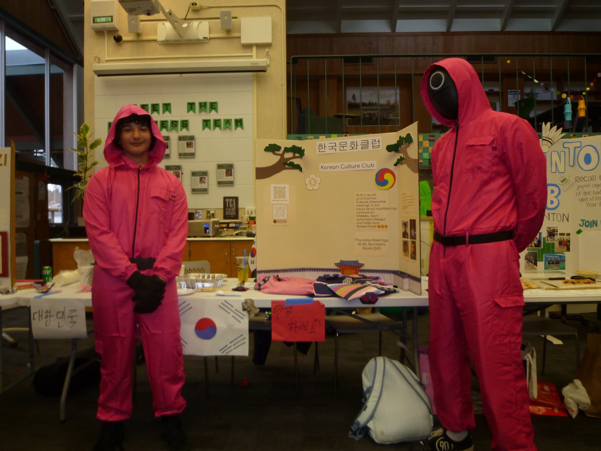 Two students in pink jumpsuits beside the Korean Culture Club's display board and a Korean flag.